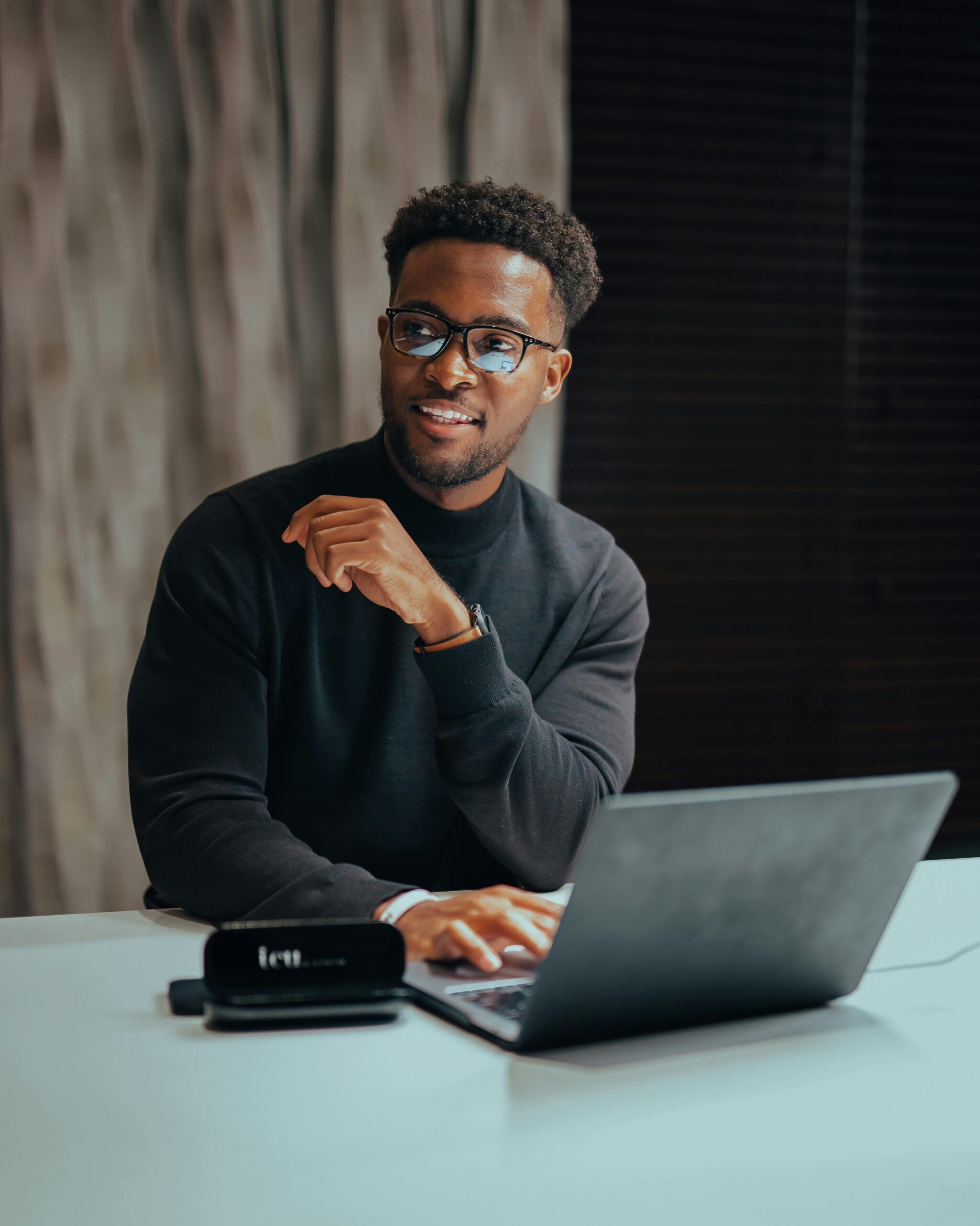 Cheerful man with glasses working on laptop at a table indoors, emanating a professional and modern workspace vibe.